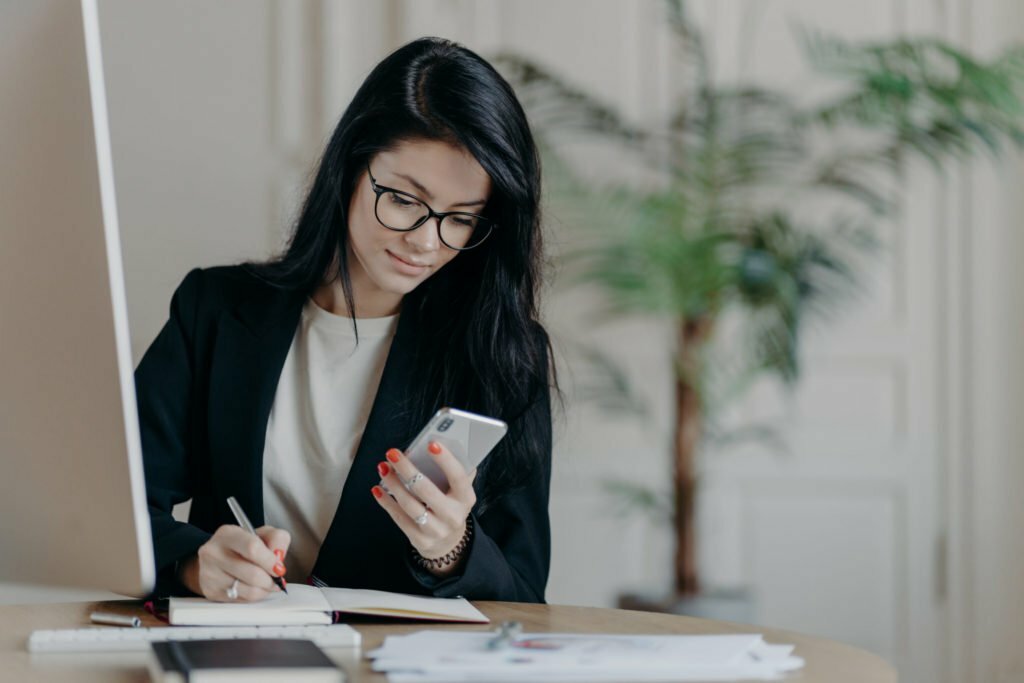 Woman with dark hair, concentrated into mobile phone, searches necessary information in internet